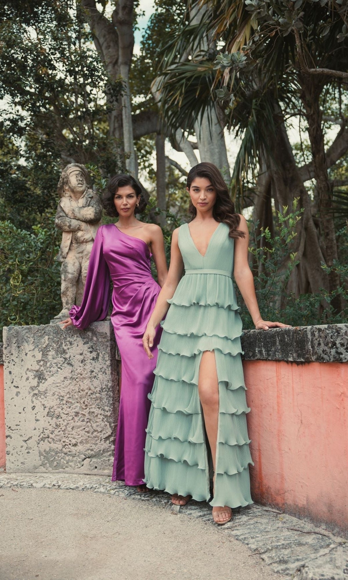 Two women pose outdoors; one wears the Mac Duggal 26588 one-sleeve long formal dress in purple, while the other wears a sleeveless mint green ruffled dress with a front slit. Trees and a statue appear in the background.